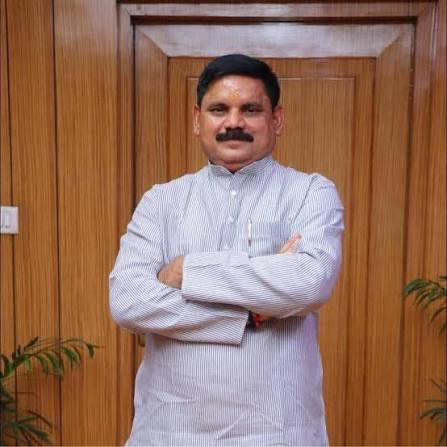 Satish Chandra Dubey standing with arms folded, wearing a pale striped kurta, in front of a wooden door