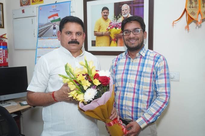 Satish Chandra Dubey holding a bouquet of flowers beside a smiling man in a checked shirt in an office