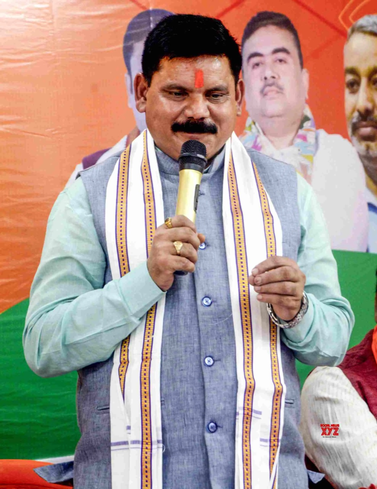 Satish Chandra Dubey speaking into a handheld microphone, wearing a grey waistcoat and an embroidered white stole