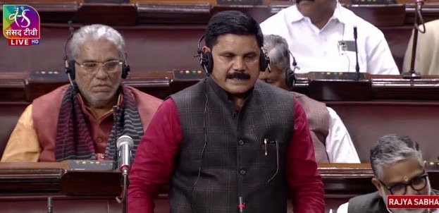 Satish Chandra Dubey standing at his seat in the Rajya Sabha, wearing a dark waistcoat and earpiece, speaking into the desk microphone